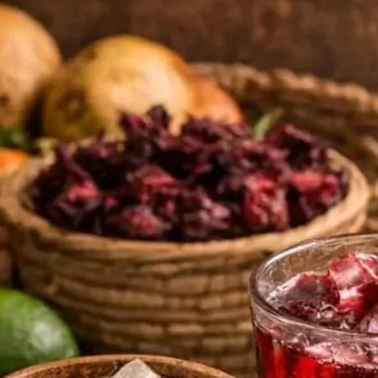 Fleurs d'hibiscus séchées dans un panier tressé, à côté d'un verre de bissap glacé